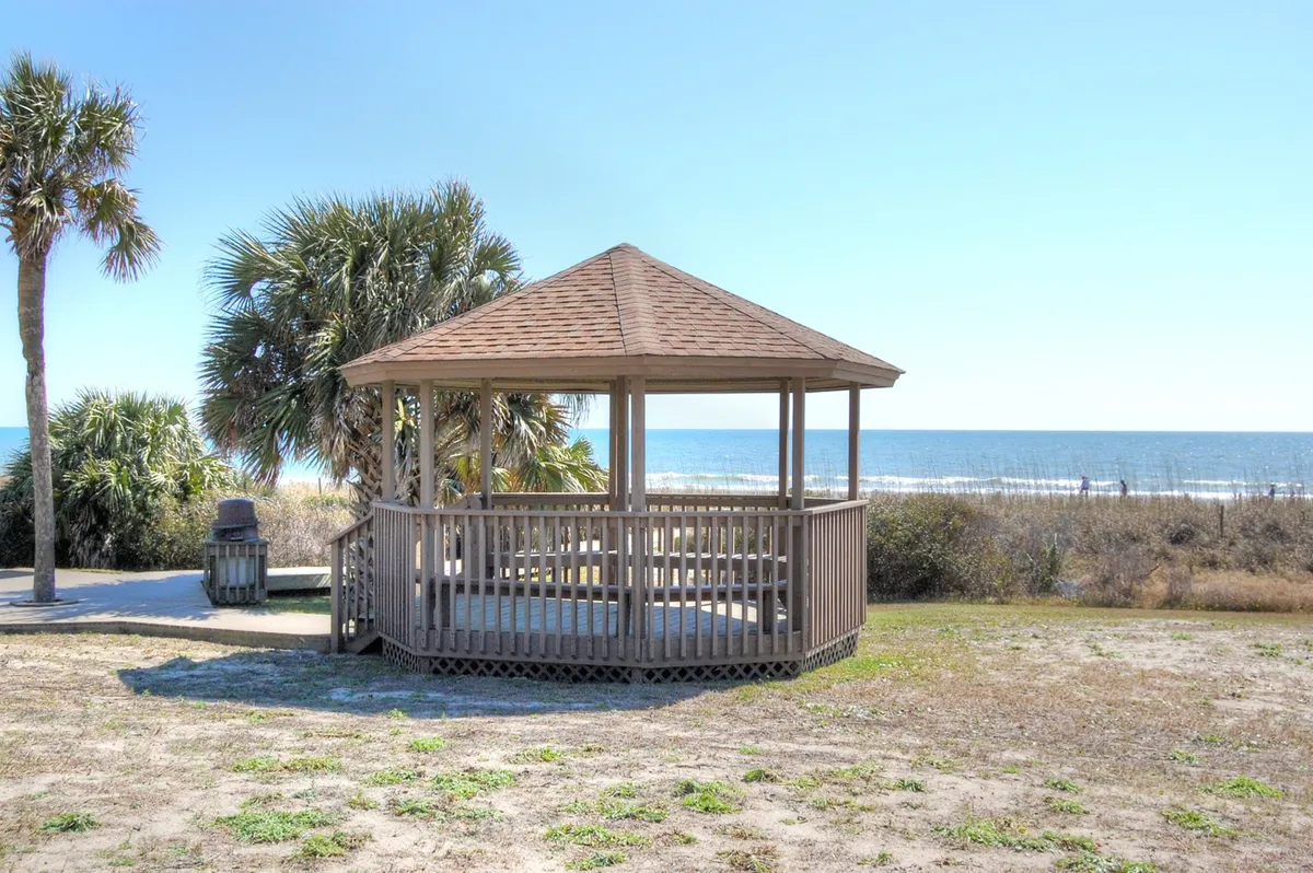 A tranquil wooden gazebo overlooking the ocean, surrounded by palm trees. The inviting structure is set against a backdrop of blue water and sky, offering ample space for small groups to relax and socialize while enjoying the coastal view.