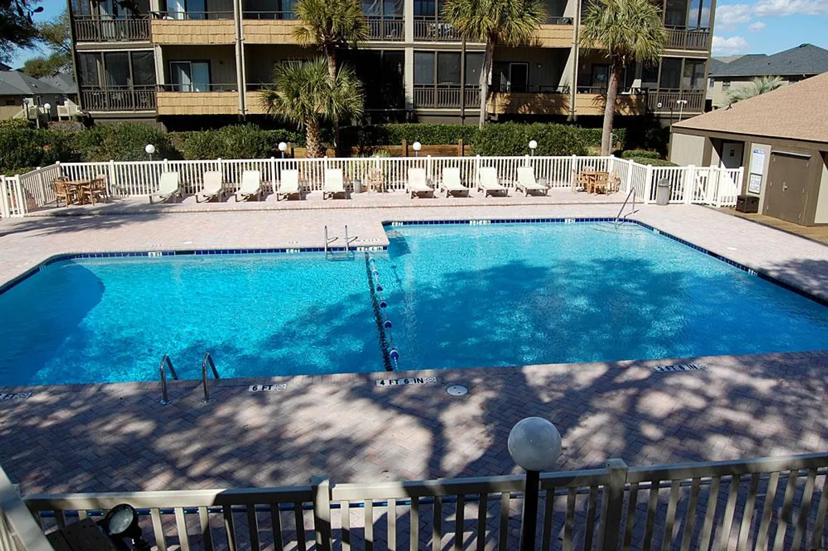 A sparkling outdoor swimming pool surrounded by lounge chairs, palm trees, and nearby resort buildings. The scene is lively and welcoming, suggesting a resort atmosphere ideal for swimming, sunbathing, and gathering with friends or family.