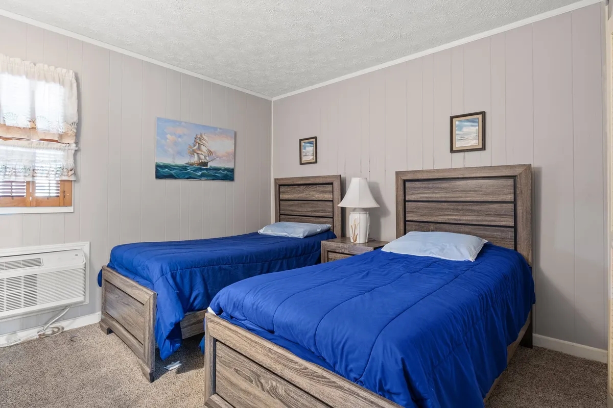 A peaceful second bedroom featuring two twin beds with blue bedding, a small bedside lamp, wall art depicting a sailing ship, and a window with sheer curtains. The overall mood is calm and welcoming, perfect for children or friends to unwind and share stories.