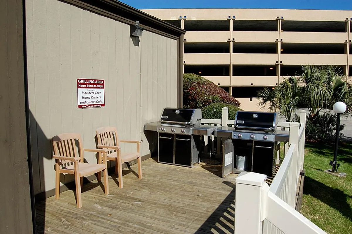 An outdoor grilling area featuring two wooden chairs and palm trees, adjacent to a parking structure. The space is designed for barbecuing, socializing, and relaxing in the sun, suggesting informal group gatherings and shared meals.