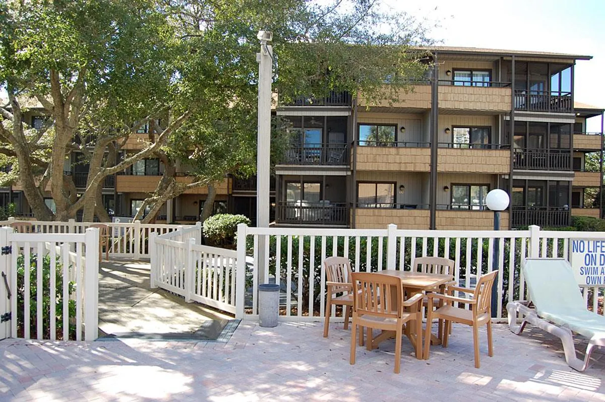 A casual outdoor setting featuring wooden chairs and a table, with apartment balconies and lush trees in the background. The space feels perfect for social gatherings, enjoying the sunshine, or taking in tranquil afternoons.