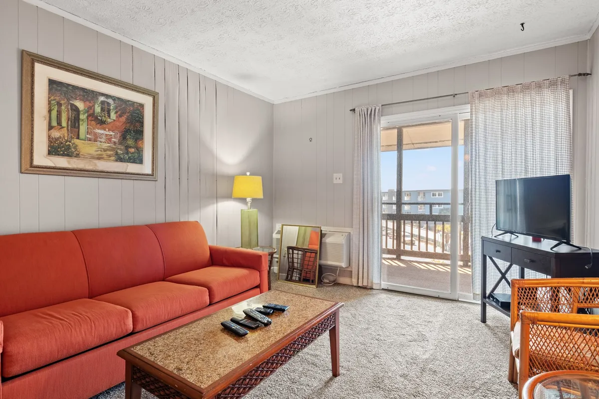 A comfortable interior view with an orange sofa (including a Queen sleeper), wooden coffee table, TV, lamp, and a balcony door that lets in sunlight. The space looks ideal for movie nights, family gatherings, or unwinding after a day of adventure.