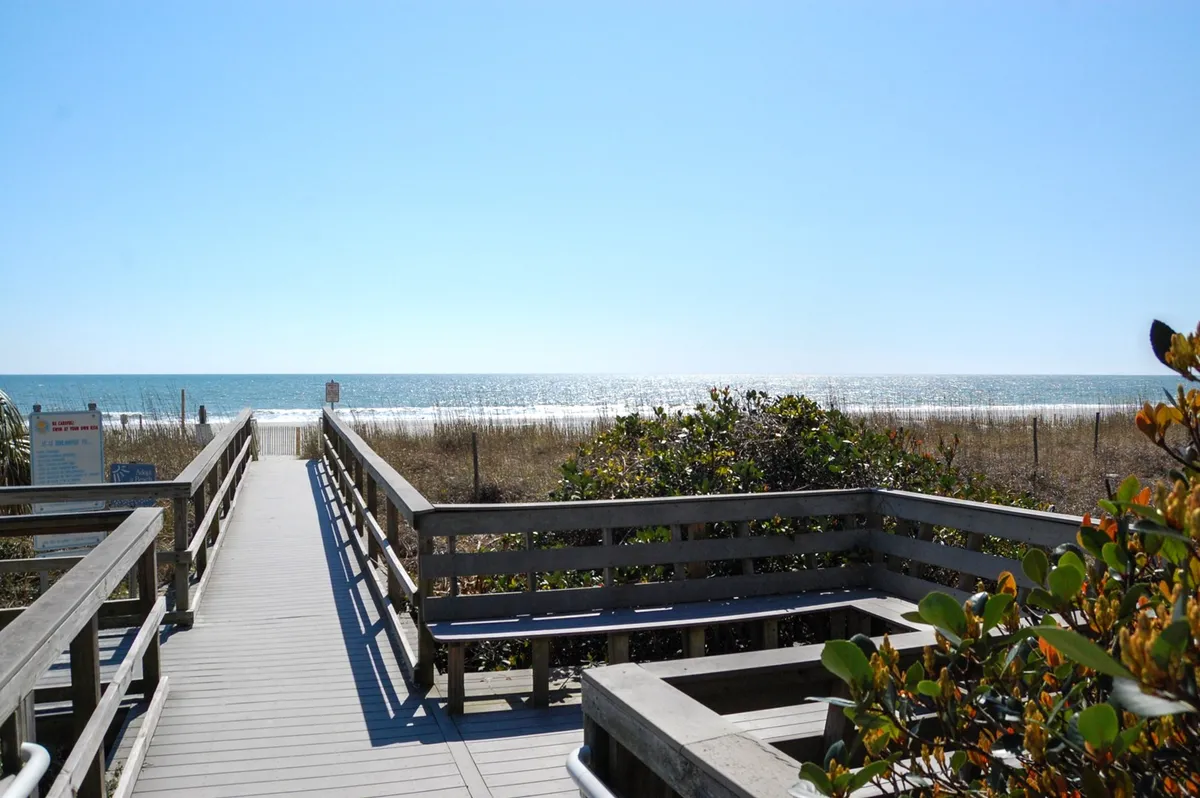 A picturesque view of a wooden boardwalk stretching toward the ocean, flanked by dunes and lush greenery. The sun hovers low, evoking a relaxed, romantic mood perfect for morning strolls by the beach.