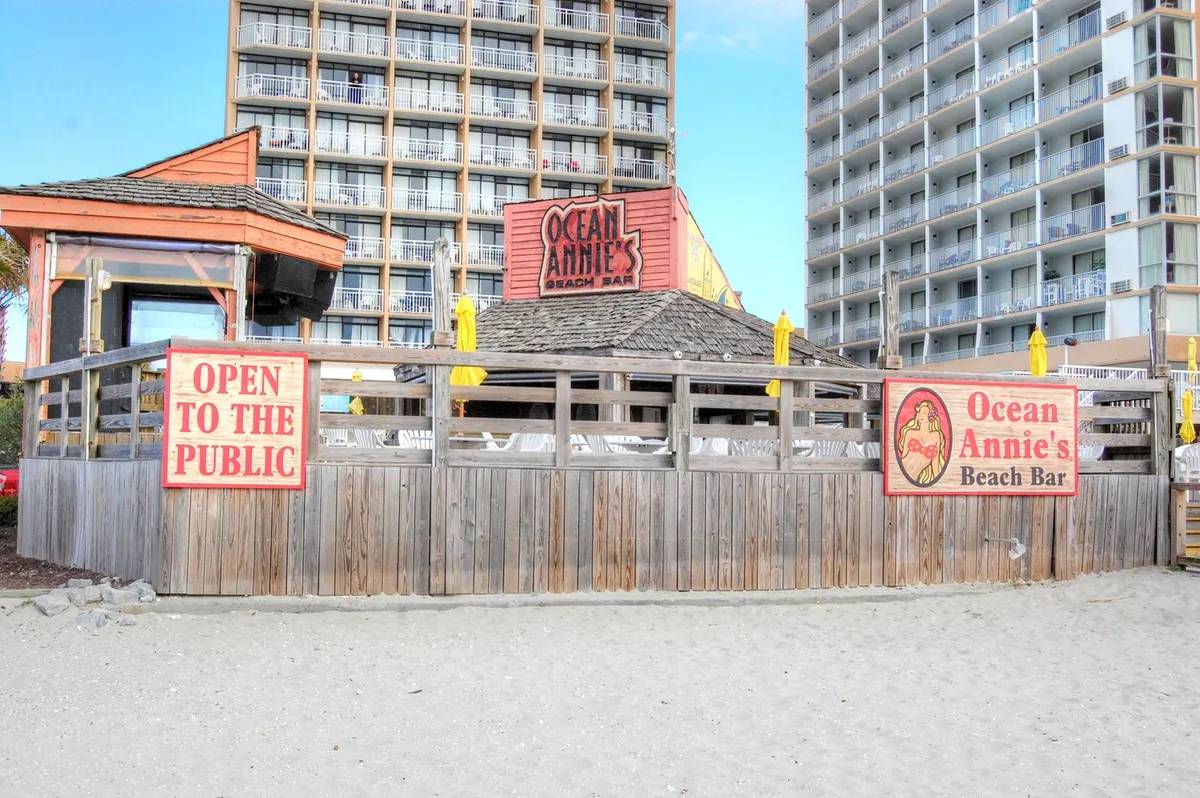 A colorful exterior shot of Ocean Annie's Beach Bar, featuring its cheerful signage, a bustling beachfront bar structure, and high-rise buildings in the background. The scene feels lively and social, suggesting music, drinks, and family-friendly energy just across the street.