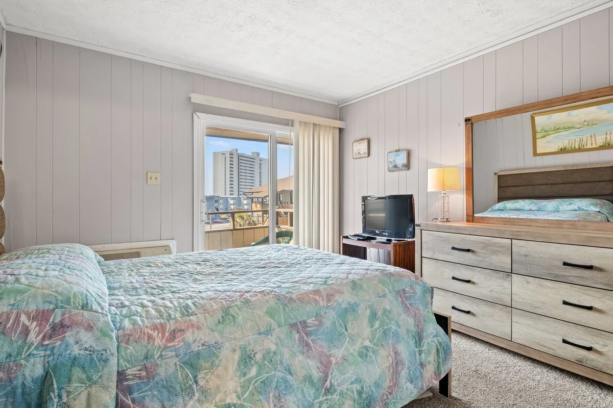 A bright, relaxed bedroom scene with a comfortable bed, dresser, window with natural light, and a wall-mounted TV. The room exudes comfort, making it ideal for unwinding after a day of adventures, with ample space for storing personal belongings.