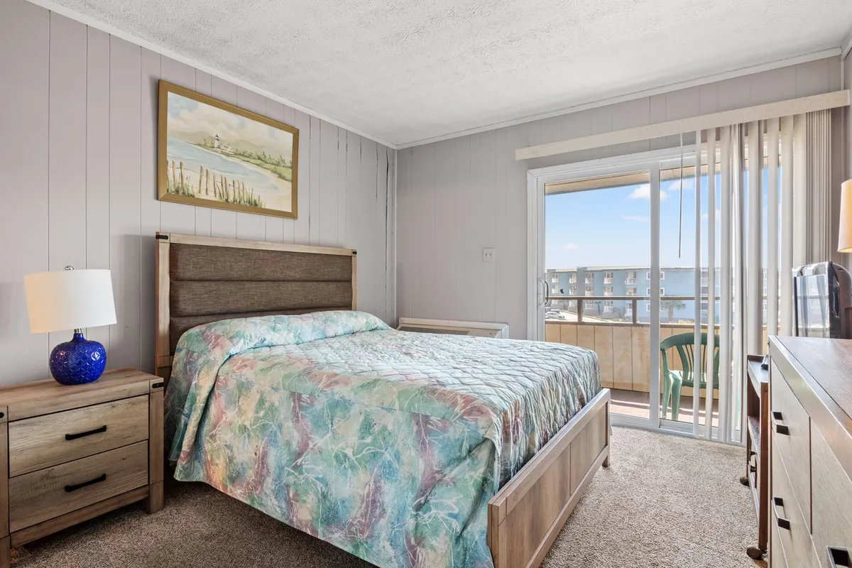 A cozy primary bedroom featuring a plush Queen bed, nightstand with lamp, a sliding glass door that opens to a balcony, and tasteful wall art. The soft linens and gentle natural light create a tranquil, restful atmosphere, with space for relaxation or enjoying a morning view from bed.