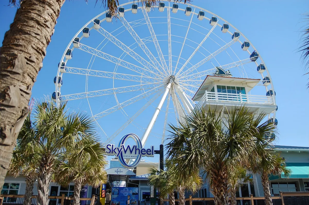 A dynamic shot of Myrtle Beach's SkyWheel, with palm trees and a ticket booth in the foreground. The iconic Ferris wheel rises above, promising fun and panoramic views—a quintessential family activity and one of the exclusive Free Fun Program attractions.